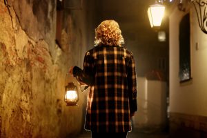 Beautiful blonde female detective with vintage lantern in retro coat walking in old street